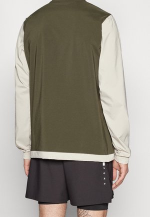 Veste verte et beige avec col montant, poignets élastiques et tissu léger, vue de dos. Portée avec un short foncé.