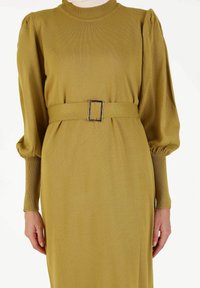Vestido de punto acanalado verde oliva, con cintura ajustada y un cinturón de tela ancho que presenta una hebilla metálica rectangular. Mangas largas y abullonadas.