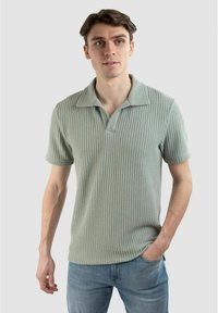 Pikétröja i ljusgrön färg, textil med strukturerad yta och ribbat mönster, korta ärmar och spetsig krage, tillsammans med blå jeans.