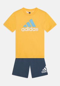 Keltainen puuvillapaita, jossa on sininen ja valkoinen Adidas-logo, yhdistettynä tummansinisiin sortseihin, joissa on valkoinen Adidas-logo lahkeessa.