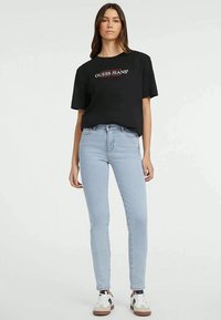 Črna majica s kratkimi rokavi z belim in rdečim napisom "GUESS JEANS", v kombinaciji s svetlo modrimi skinny džinsom. Obutev ima belo osnovo z rjavimi poudarki.