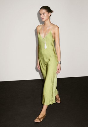 Long green satin slip dress with thin straps, featuring a deep neckline and flowing design. Worn with brown flat sandals and layered necklace.