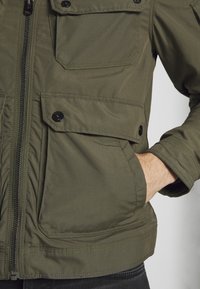 Olivgrön isolerad jacka i ett slätt tyg. Har dragkedja, flera fickor och kontraststickning längs fållarna.