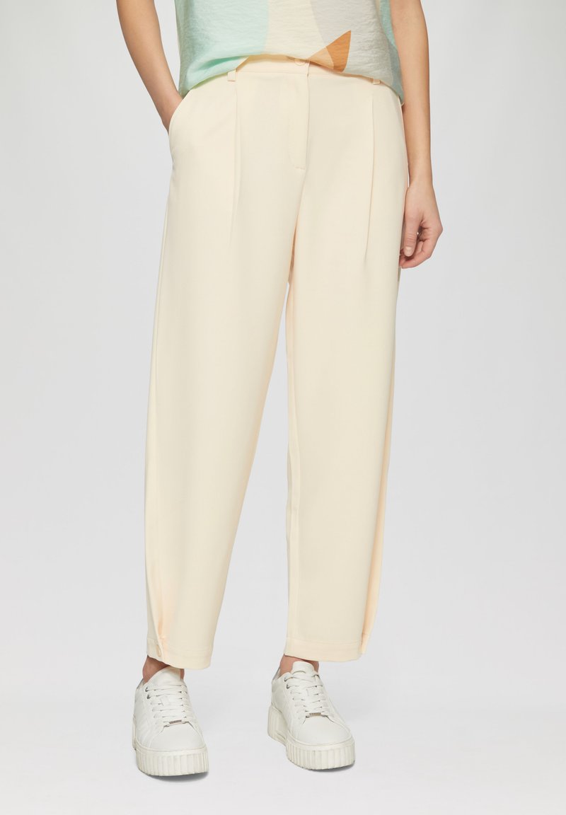 Pantalons ajustés couleur crème avec une coupe ample, dotés de plis à l'avant, de poches latérales et d'une texture lisse. Associés à des baskets blanches.