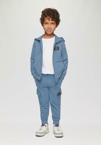 Blauer Baumwollhoodie mit Reißverschluss und Taschen, kombiniert mit passenden Jogginghosen mit Cargo-Taschen und einem schwarzen Logo-Patch. Weiße Sneaker.