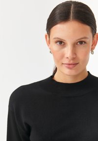 Fekete kötött pulóver magas bordás nyakkal, sima textúrával, egy sima fehér háttér előtt.