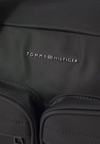 Svart stoffeveske med stiktede detaljer og to framlommer. Viser preget sølv "TOMMY HILFIGER"-logo i midten.