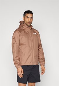 Brun vandtæt jakke med hætte, der har elastiske manchetter og lynlås. Logo på brystet. Båret over mørke shorts. Glat tekstur.