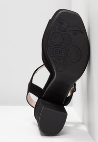 Sandale en daim noir avec un talon épais, comportant deux sangles traversant le pied et une semelle en caoutchouc noir texturé avec des détails en relief.