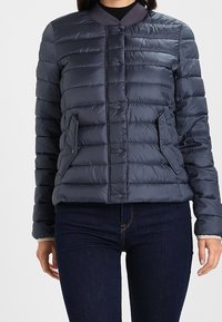 Donna che indossa un giubbotto trapuntato blu navy con bottoni a scatto e tasche laterali, abbinato a jeans in denim blu scuro.