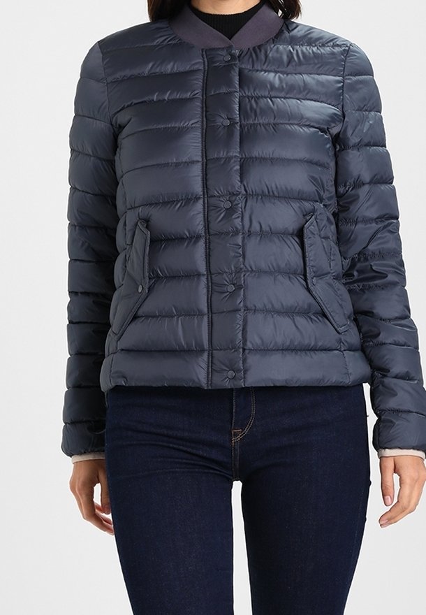 Donna che indossa un giubbotto trapuntato blu navy con bottoni a scatto e tasche laterali, abbinato a jeans in denim blu scuro.