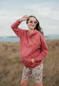 Femme souriante en plein air portant des lunettes de soleil, un sweat à capuche rouge et un short fleuri, une main dans la poche et l'autre touchant ses lunettes de soleil.