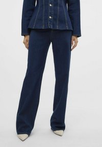 Temno modri jeans hlače s ravnim krojem, z srednje visokim pasom in diskretnim oranžnim šivom. Uporabljene z svetlo obarvanimi koničastimi čevlji.