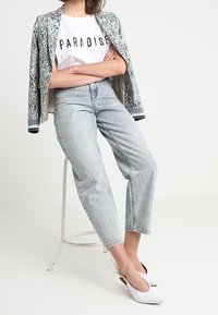 Weißes Grafik-T-Shirt mit dem Text "PARADISE", leichte blau gemusterte Jacke und hellblaue gerade Jeans, kombiniert mit weißen Absatzmules.