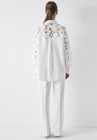 White blouse with floral lace panel on sleeves and back, paired with white fitted trousers. Smooth texture, collar detail visible.