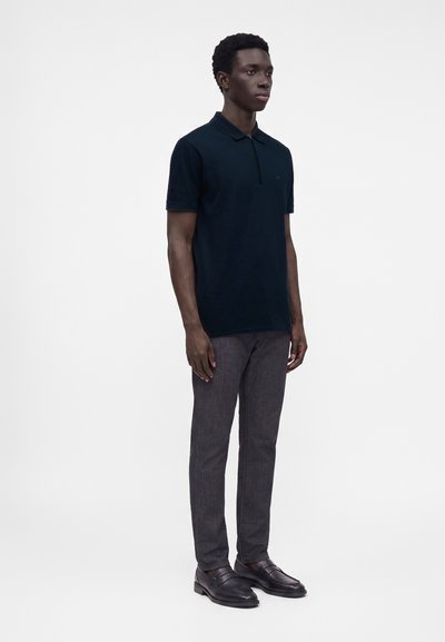 Jeune homme debout portant un polo noir à manches courtes, un pantalon gris foncé et des chaussures habillées noires sur un fond blanc uni.
