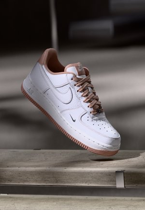 Biały i jasnoróżowy sneaker Nike Air Force 1 zawieszony nad betonową ławką na neutralnym szarym tle.