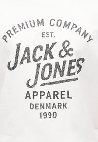 Κείμενο λογότυπου Jack & Jones σε λευκό ύφασμα με τη φράση «Premium Company Est. Apparel Denmark 1990» σε φθαρμένη μαύρη γραμματοσειρά.