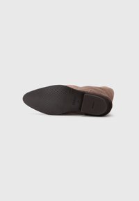 Bottines en suède marron avec un bout pointu ; semelle en caoutchouc noir texturé ; marque minimaliste sur la semelle intérieure ; pas de matériel ou d'accents visibles.