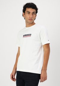 Valge puuvillane t-särk lühikeste varrukatega, mille esiküljel on nelinurkne Tommy Hilfiger logo meresinistes ja punastes triipudes.