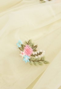 Gesticktes Blumenmuster mit rosa, blauen und grünen Fäden auf transparentem, hellgelbem Stoff mit feiner Textur und Drapierung.