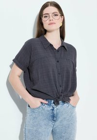 Camisa gris de manga corta con un patrón a cuadros y botones al frente, atada a la cintura. Se lleva con unos vaqueros de tiro alto, de color azul claro y desgastados.