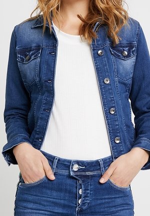 Femme portant une veste en jean bleue et un jean avec un t-shirt blanc côtelé, mains dans les poches avant, cheveux lâchés avec de douces ondulations.