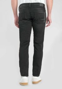 Vaqueros de mezclilla negros con un ajuste Slim, que presentan detalles de costura sutiles y un bolsillo con zipper en la parte posterior. Llevados con zapatillas blancas.