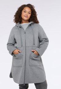 Cappotto grigio in misto lana con zip frontale, cappuccio e due tasche anteriori. Presenta una vestibilità comoda e un lieve orlo a forma di A. Tessuto morbido.