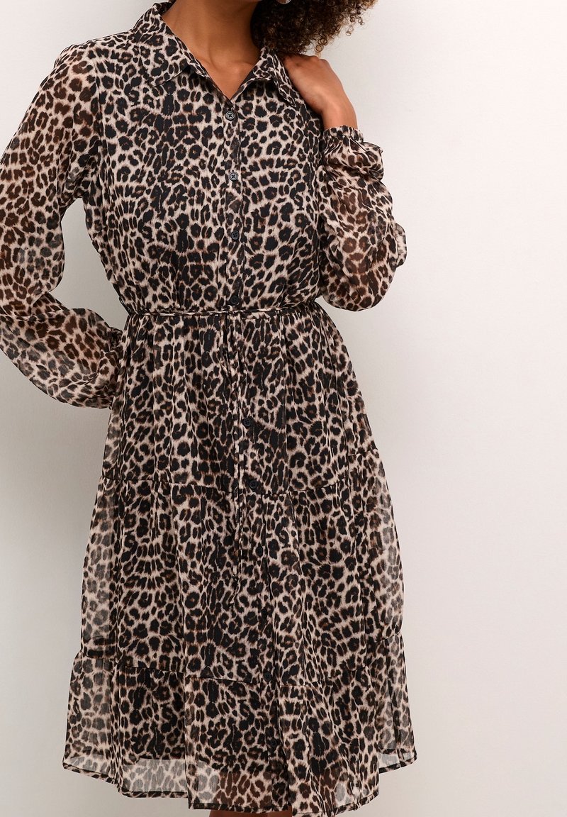 Femme portant une robe à manches longues boutonnée, imprimé léopard, avec une taille froncée et une jupe à volants, debout devant un fond clair uni.