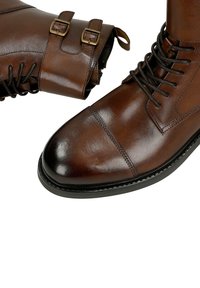 Stivali in pelle marrone con finitura lucida, chiusura frontale con lacci e doppi dettagli con fibbia alla caviglia. Suola robusta con dettagli di cucitura.