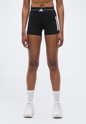 Athlète debout portant un short Adidas noir à bandes blanches, des chaussettes blanches montantes et des baskets Adidas blanches sur un fond uni.