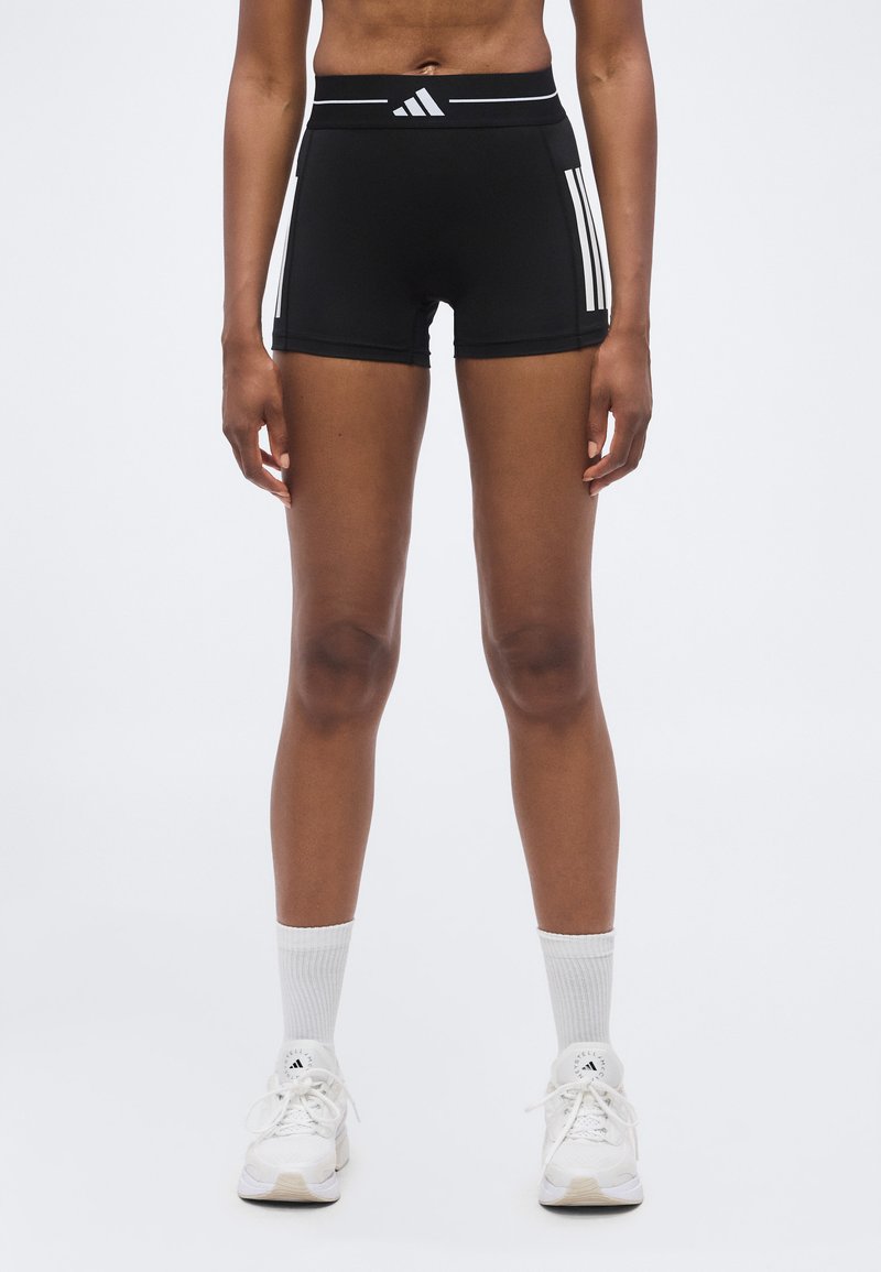 Athlète debout portant un short Adidas noir à bandes blanches, des chaussettes blanches montantes et des baskets Adidas blanches sur un fond uni.