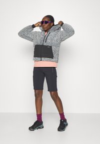 Veste en polaire zippée grise avec une poche avant noire, accompagnée de shorts noirs, de chaussettes violettes et de chaussures de sport noires et teal.
