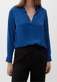 Camicia a maniche lunghe blu con scollo a V e colletto, caratterizzata da una texture stropicciata e una vestibilità rilassata, abbinata a pantaloni neri.