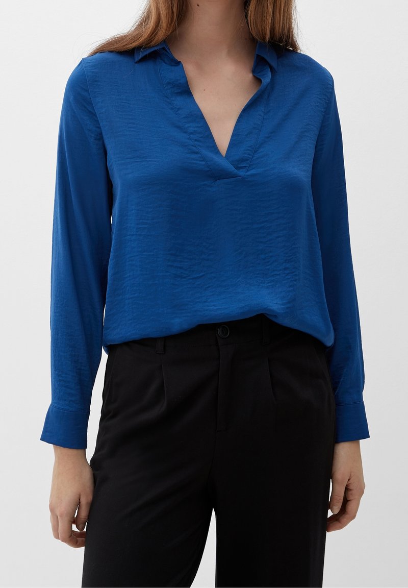Camicia a maniche lunghe blu con scollo a V e colletto, caratterizzata da una texture stropicciata e una vestibilità rilassata, abbinata a pantaloni neri.