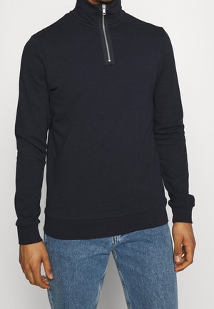 Homme portant un pull noir à manches longues avec fermeture éclair au quart et un jean bleu, debout devant un fond clair uni.