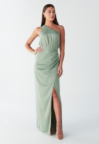 Lange, asymmetrische jurk in zachtgroene stof met een geplooide taille. Voorzien van een hoge split aan de linkerkant en een gladde textuur.
