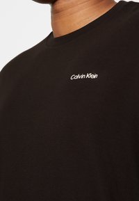 Sort bomulds T-shirt med en klassisk rund hals, prydet med et hvidt "Calvin Klein" logo på venstre side af brystet. Glat tekstur.