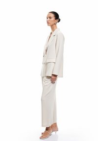 Beige dubbelzijdige blazer met notch revers, gecombineerd met bijpassende brede broek. Het model draagt open teen sandalen met banden.