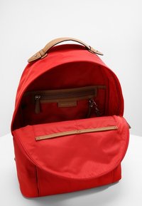 Rød nylon rygsæk med et sandfarvet læderhåndtag og lynlåsdetaljer. Har et rummeligt interiør samt en ekstra lynlåslomme.