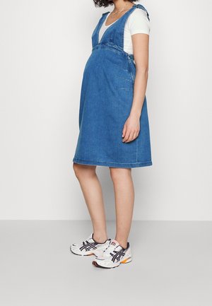 Gravid kvinde iført blå denim-forklædekjole over hvid t-shirt og hvide sneakers, stående mod en ensfarvet lys baggrund.