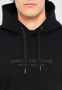 Schwarzer Baumwollhoodie mit Kapuze und Kordelzügen. Auf der Vorderseite ist der erhabene Schriftzug "ARMANI EXCHANGE MILANO / NEW YORK" zu sehen. Glatte Textur.
