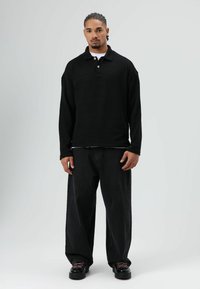 Zwarte poloshirt met lange mouwen en drie knopen, gedragen over een wit shirt, gecombineerd met losse zwarte jeans en zwarte veterschoenen.