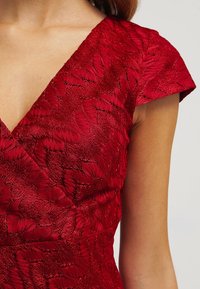 Robe rouge avec un décolleté en V et des manches courtes. Le tissu présente un motif de feuilles texturé, ajoutant de la profondeur et de l'intérêt visuel.