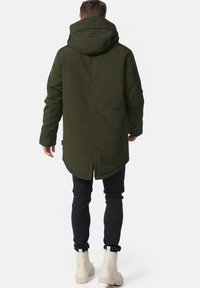 Olive green hooded parka with a longer back hem, textured fabric, and side slits, paired with dark slim-fit pants and light-colored boots.