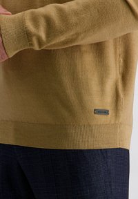 Senfgelber strickpullover mit gerippten Bündchen und Saum, ausgestattet mit einem kleinen Logo-Etikett auf der unteren Seite. Die Textur erscheint weich und glatt.