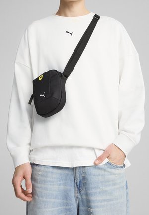 Personne portant un sweat-shirt Puma blanc et un jean bleu clair avec un sac bandoulière Puma et Ferrari noir porté en travers de la poitrine.