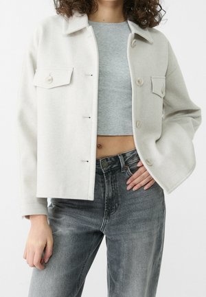 Person wearing a light beige button-up jacket over a gray crop top and faded black jeans, left hand on hip, facing forward.