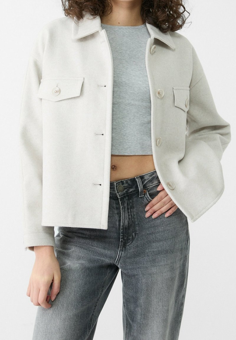 Personne portant une veste boutonnée beige clair sur un crop top gris et un jean noir délavé, main gauche sur la hanche, faisant face vers l’avant.